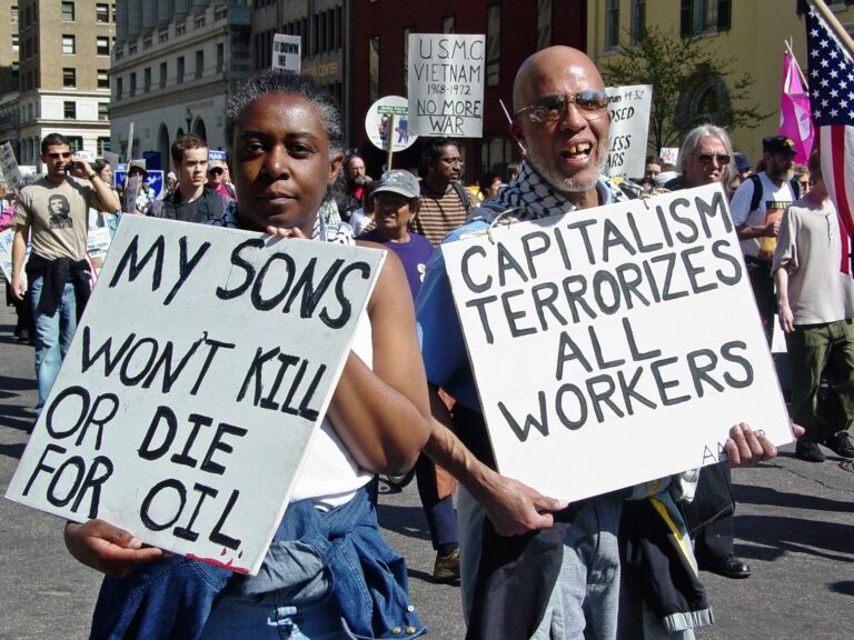 A Black woman holds a sign that reads "my sons won't kill or die for oil" and a Black man holds a sign reading "Capitalism terrorizes all workers."