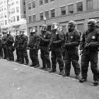 A black and white photo of police officers wearing riot gear standing in front of buildings.