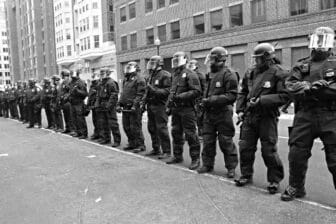 A black and white photo of police officers wearing riot gear standing in front of buildings.