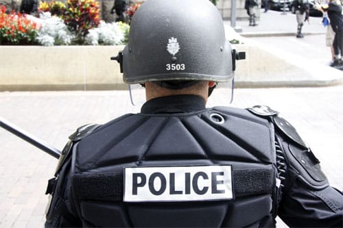 Photo of a police officer from behind. He is wearing a thick vest that reads "POLICE" and a helmet.