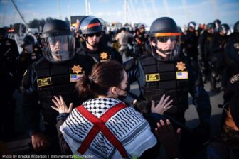 A woman wearing a keffiyeh has her hands up as a wall of police officers approaches.
