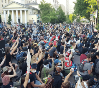 Very large group of protestors filling a street with their hands raised in the air. There is also a light yellow building with pillars in the back.