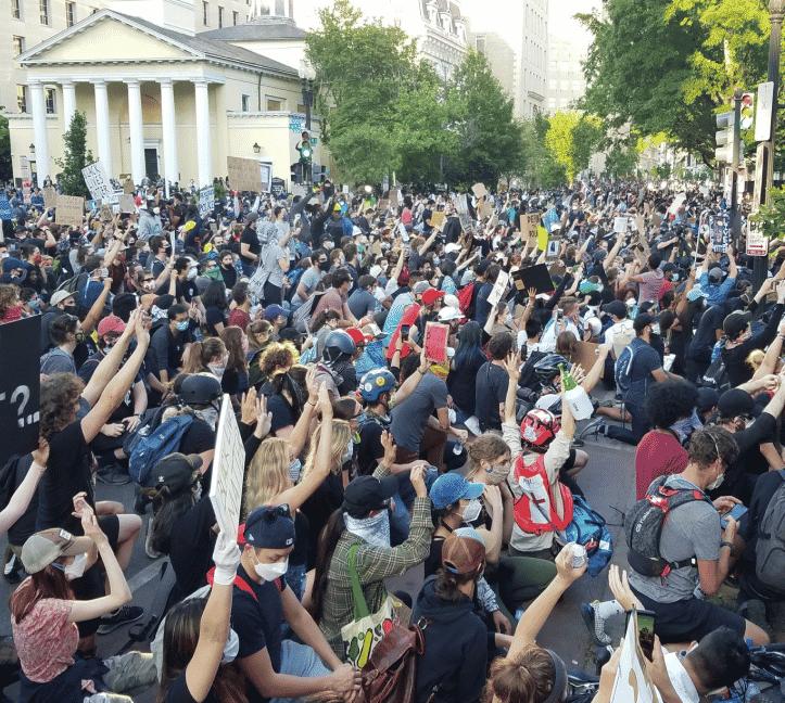 Very large group of protestors filling a street with their hands raised in the air. There is also a light yellow building with pillars in the back.